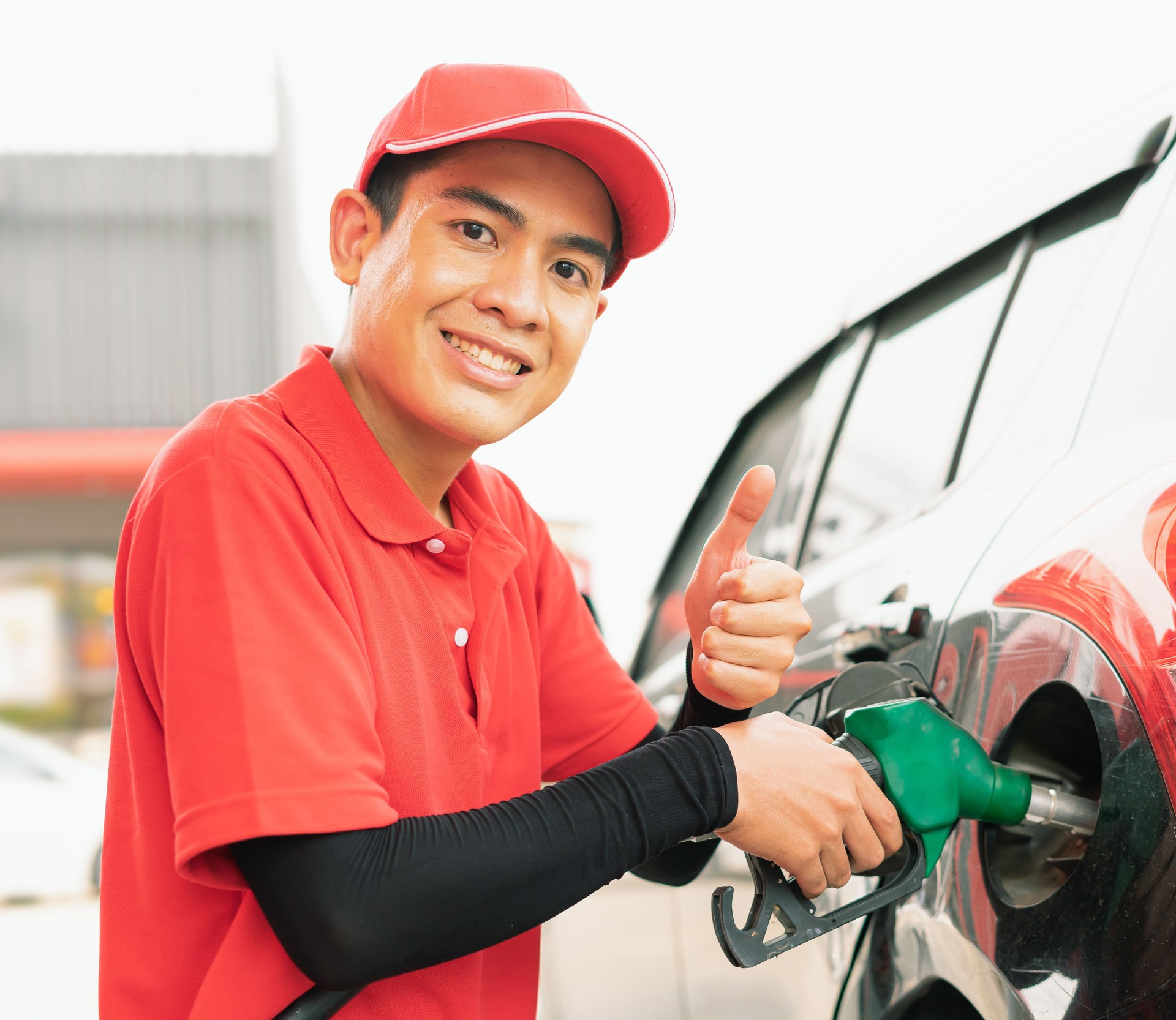 Asian gas station worker man give thumbs up in one hand and holding green fuel nozzle into one hand for filling high energy power fuel into black auto car tank in petrol station, commercial service for benzine, diesel, gasohol, gasoline